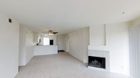 This single-floor space features an open-concept layout that seamlessly connects the living area to the kitchen. The living room is a generously sized, light-filled space with neutral wall tones, smooth white ceilings, and plush wall-to-wall carpeting that adds warmth underfoot. A brick-surround fireplace serves as a natural focal point along one wall, offering both visual interest and functional comfort. A ceiling fan is centrally positioned overhead, providing efficient air circulation throughout the space. The kitchen is partially open to the living area, divided by a raised breakfast bar that provides a convenient casual dining or prep surface. The kitchen features dark appliances, including a refrigerator, and is accessible via the main living space. Natural light enters through a window visible near the kitchen area, contributing to the bright and airy atmosphere throughout. A hallway leads to additional areas of the home. The overall aesthetic is clean and neutral, with a simple, functional design that offers a versatile canvas for personalization.