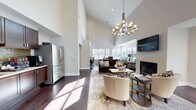 Located at 800 Runaway Bay Lane in Mount Pleasant, South Carolina, this single-floor community space is designed to impress with its dramatic vaulted ceilings and open-concept layout. The main living area serves as the heart of the space, anchored by a tile-surround fireplace with a mounted flat-screen television above, and illuminated by a striking multi-arm chandelier with candle-style shades. Rich dark hardwood flooring runs throughout, complementing the neutral gray-and-white color palette on the walls. Comfortable seating arrangements, including plush sofas and accent chairs, are arranged around a central coffee table atop a patterned area rug, creating an inviting lounge atmosphere. Natural light pours in through an array of windows, including elevated clerestory windows that enhance the sense of height and openness. French doors provide access to an outdoor area. The kitchenette is outfitted with dark wood cabinetry, a mosaic tile backsplash, stainless steel appliances including a full-size refrigerator, and ample counter space. Adjacent to the main lounge is a sun-drenched dining area with a dark wood high-top table surrounded by upholstered chairs, alongside a cozy seating nook. A dedicated business center features a built-in desk with dual workstations, pendant lighting, and a distinctive light blue accent wall, providing a functional and focused workspace within the broader communal setting.