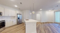 This well-appointed single-floor residence showcases a clean, contemporary design throughout. The heart of the home is a bright, open kitchen featuring crisp white cabinetry, a decorative tile backsplash, and a full suite of stainless steel appliances, including a side-by-side refrigerator with water dispenser and a built-in microwave and range. A central island with a white quartz countertop and an integrated sink serves as both a functional workspace and a social hub, accented by stylish pendant lighting overhead. Warm-toned luxury vinyl plank flooring runs throughout the kitchen and living areas, lending the space a cohesive, modern feel. The bathrooms are thoughtfully finished with white quartz vanities, modern brushed hardware, wood-look flooring, and striking marble-patterned tile surrounds encasing the soaking tub and shower combinations — adding an elegant, spa-like quality. The bedrooms are finished with soft, neutral carpeting and include ceiling fans for year-round comfort. Natural light filters in through window treatments, brightening the spacious, freshly painted white walls. Recessed lighting throughout ensures every area is well-illuminated, creating an inviting and functional living environment.