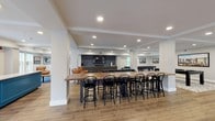 This single-floor community space is thoughtfully designed to accommodate a variety of social and functional activities within an open, modern layout. The interior is finished with warm hardwood-style flooring throughout, crisp white walls, and recessed lighting that creates a bright, welcoming atmosphere.

The main gathering area features a comfortable lounge zone anchored by a blue-toned area rug, mid-century modern seating including leather accent chairs, and a wall-mounted television set into a dark-cabinetry media nook. Large windows along multiple walls flood the space with natural light and provide views of the surrounding greenery and outdoor patio areas.

A generously sized communal kitchen serves as a central hub, outfitted with stainless steel appliances including a refrigerator, dishwasher, range, and a statement island range hood. Dark cabinetry contrasts cleanly with light quartz countertops, and a window above the sink looks directly onto an outdoor seating area.

Multiple dining configurations are available, including a long bar-height communal table with classic black bentwood stools and a separate standard-height dining table seating up to eight. A game area with a pool table and shuffleboard rounds out the space, making this a well-equipped, versatile community amenity.