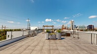 This rooftop terrace offers a well-appointed outdoor experience atop a modern building, with large-format porcelain tile flooring extending across a generously sized deck. The terrace is divided into distinct functional zones, beginning with a central gathering area anchored by a wooden pergola and clustered cylindrical planters filled with lush greenery, providing both shade and a naturalistic focal point. Seating areas are positioned throughout the deck to take advantage of the panoramic surroundings.

A section of vibrant artificial turf introduces a lawn-like element to the rooftop, complemented by tall dark-toned rectangular planters and additional seating that face directly toward the Philadelphia city skyline—a striking backdrop visible from this vantage point. The skyline view captures a broad sweep of the urban horizon under open sky.

A clean, modern rooftop access structure with glass double doors serves as the entry point, clad in white panel siding and marked with building branding. Metal railings border the perimeter on all sides, ensuring safety while maintaining unobstructed sightlines to the surrounding industrial and urban landscape. The overall ambiance is open, contemporary, and city-connected.