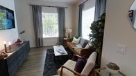 This single-floor space showcases a well-appointed living room that blends modern aesthetics with mid-century design sensibilities. The room is anchored by large windows dressed with elegant grey curtains and white horizontal blinds, allowing an abundance of natural light to fill the space while offering a view of lush greenery outside.

The living area features warm hardwood flooring that complements a rich cognac leather sofa adorned with an assortment of decorative pillows in neutral and teal tones. A pair of cream-upholstered mid-century armchairs with wood frames adds a classic touch, while a geometric-patterned area rug in deep charcoal grounds the seating arrangement. At the center, a rectangular coffee table with a marble-top surface and wooden legs serves as both a functional and decorative focal point.

Thoughtful styling details elevate the space, including a tall fiddle-leaf fig tree that introduces a vibrant natural element, a slender table lamp with a warm glow, and carefully placed accent décor. The neutral wall tones provide a clean backdrop that ties the entire room together, creating an inviting, balanced atmosphere that is both contemporary and comfortable.