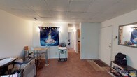 This single-floor space features an open layout with a carpeted living area serving as the central gathering space. The room is finished with a drop tile ceiling and light-colored walls, giving the space a simple, functional character. A small dining table and chairs occupy one corner, while a hallway leads toward additional rooms at the rear of the unit. The kitchen, visible from the main living area, includes wood-toned cabinetry and appears compact yet practical. Natural light is supplemented by overhead and wall-mounted lighting throughout. A doormat near what appears to be an entry point indicates a transition between the carpeted interior and a hard surface flooring area near the entrance. The overall layout is straightforward, with clear sightlines from the living area to the kitchen and hallway, making the space feel connected despite its modest scale. The neutral tones of the walls and flooring provide a versatile backdrop, and the drop ceiling is consistent throughout the visible areas of the space.