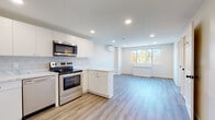 This single-floor space showcases a clean, modern aesthetic throughout, highlighted by light-toned luxury vinyl plank flooring that flows seamlessly across the main living areas. The open-concept kitchen is a standout feature, equipped with stainless steel appliances including a refrigerator, electric range, over-the-range microwave, and dishwasher. Crisp white cabinetry extends from floor to ceiling, complemented by a marble-look quartz countertop and a decorative hexagonal tile backsplash. A peninsula counter provides a natural division between the kitchen and the open living area, enhancing both functionality and flow. Recessed lighting throughout ensures consistent, bright illumination across the space. A wall-mounted mini-split unit offers climate control in the living area, which benefits from a large window framing lush, tree-lined outdoor views. The bedroom features soft carpeting, a wide sliding-panel closet, and a generous window that fills the room with natural light and scenic greenery. The full bathroom is finished with large-format neutral wall tiles surrounding a soaking tub with an overhead shower, paired with a white vanity and tile flooring. Overall, the space presents a fresh, well-appointed interior with a cohesive neutral palette and thoughtful modern finishes throughout.