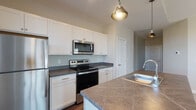 Located at 7325 Liberty Summit Dr in Powell, Ohio, this single-floor residence offers a clean, contemporary design with functional living spaces. The kitchen is a standout feature, showcasing crisp white shaker-style cabinetry that extends to the ceiling for ample storage. Dark stone countertops provide a striking contrast against the white cabinets, while a full suite of stainless steel appliances — including a refrigerator, electric range, and over-the-range microwave — delivers a polished, modern look. A kitchen island with a stainless steel sink adds both workspace and convenience. Stylish pendant lighting with warm-toned metal shades hangs above the island, enhancing the kitchen's inviting ambiance. The light neutral wall tones and wood-style flooring contribute to a bright, cohesive aesthetic throughout. The thoughtful layout promotes an open flow between the kitchen and adjoining spaces, making the home well-suited for both everyday living and entertaining. The overall design balances modern finishes with a warm, approachable character, creating a comfortable and well-appointed living environment.