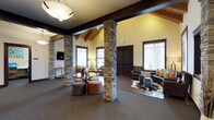 This single-floor space offers an impressive and inviting atmosphere defined by its striking architectural details and thoughtful design. Soaring vaulted ceilings with exposed wood beam accents and tongue-and-groove paneling draw the eye upward, creating an airy, lodge-inspired ambiance throughout. Prominent stone columns anchor the open floor plan, adding a rugged, natural texture that complements the warm, earthy color palette of the interior.

The space is divided into distinct yet connected seating areas, each designed to encourage conversation and relaxation. One area features a glass-top coffee table surrounded by cushioned stools in warm terracotta and neutral tones, while an adjacent lounge zone is furnished with rich leather seating arranged over a cowhide-style area rug, lending a rustic yet refined character. A wall-mounted flat-screen television is positioned for easy viewing from the seating areas.

Natural light filters in generously through multiple windows fitted with blinds, brightening the neutral-toned walls and dark carpeted flooring. Modern pendant and globe light fixtures provide warm supplemental lighting. Dark-stained wood doors add a bold contrast to the light walls. The overall design blends rustic and contemporary elements, resulting in a space that feels both welcoming and well-appointed.