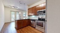 This single-floor, one-bedroom, one-bathroom unit is efficiently designed to maximize functionality within a compact footprint. The open-concept kitchen and living area serves as the heart of the unit, where rich hardwood-style flooring extends throughout. The kitchen is well-appointed with warm walnut-toned cabinetry reaching toward the ceiling, dark granite countertops, a peninsula with a double sink, stainless steel appliances including a range, built-in microwave, dishwasher, and refrigerator, and a light green glass tile backsplash. Pendant lighting above the peninsula and track lighting add layers of illumination. Multiple windows and French doors in the living area invite generous natural light and outdoor views.

A hallway connects the living space to the private bedroom, which features plush carpeting, a ceiling fan, and windows that frame greenery outside. An adjacent walk-in closet offers wire shelving for organized storage. The full bathroom includes large-format tile flooring, a tiled tub-and-shower surround, a wood-toned vanity with dark countertop, and clean, neutral finishes throughout.