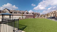 This residential community showcases a thoughtfully designed outdoor environment centered around a resort-style swimming pool with a broad brick-paver surround. Blue lounge chairs line the pool deck, offering comfortable spaces for relaxation, while umbrella-shaded tables and chairs nearby provide additional seating for outdoor dining or socializing. The pool area is framed by mature trees and neatly maintained landscaping, including trimmed shrubs and garden beds that add greenery and visual structure throughout.

Adjacent courtyard areas feature decorative brick-paver walkways connecting various seating zones, creating a cohesive and inviting communal atmosphere. A spacious open lawn area, finished with lush artificial turf, is enclosed by black metal fencing, providing a clean and versatile green space. Additional amenities include outdoor benches, metal picnic-style tables, and dedicated lighting poles for evening use.

The surrounding multi-story buildings are constructed with classic brick exteriors, gabled rooflines, and balconies that overlook the community grounds. The architectural style is consistent throughout, blending traditional residential design with modern community-focused amenities. The overall ambiance is open, well-maintained, and designed to encourage outdoor activity and community engagement across the property.