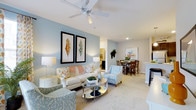 This single-floor residence showcases an open-concept design that integrates the living, dining, and kitchen areas into one cohesive, flowing space. The living area serves as the heart of the home, featuring plush carpeted flooring, a ceiling fan, and a soft blue accent wall adorned with botanical framed artwork. A comfortable seating arrangement surrounds a mirrored coffee table, creating an inviting atmosphere well-suited for both relaxing and entertaining.

The space transitions naturally into the dining area, which accommodates a dark wood table and chairs, lending a warm contrast to the cool, neutral color palette throughout. A glimpse of the kitchen is visible beyond, featuring light countertops and cabinetry that contribute to the overall brightness of the home.

Recessed lighting and a ceiling fan ensure the space remains well-lit and comfortable. The consistent use of soft beiges, muted blues, and warm whites throughout creates a calm, cohesive ambiance. The open layout maximizes the sense of space, making the residence feel airy and functional without visual clutter.