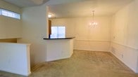 This single-floor space showcases an open, flowing layout that connects the kitchen, living, and dining areas seamlessly. A defining architectural feature is the kitchen pass-through counter with a dark contrasting countertop, which provides a visual separation between the kitchen and the adjacent living space while maintaining an open feel. The kitchen area, visible through the pass-through window, allows natural light to filter into the main living space. The living and dining areas are finished with neutral wall-to-wall carpeting that complements the warm, cream-toned walls throughout. A decorative chandelier with candelabra-style lighting serves as a focal point in the dining area, adding a touch of elegance to the space. The vaulted or raised ceiling adds a sense of vertical spaciousness, enhancing the overall openness of the layout. Clean white baseboards and trim provide crisp architectural detailing throughout. The neutral color palette of the walls and flooring creates a versatile backdrop well-suited for a variety of furnishing styles, contributing to the space's bright and welcoming ambiance.