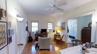 This single-floor space exudes a warm, lived-in charm with rich hardwood flooring that extends throughout, complemented by crisp white walls and classic trim work. The main living area is generously proportioned, anchored by a five-blade ceiling fan that adds both function and a casual, relaxed aesthetic. Two double-hung windows along the far wall allow natural light to fill the room, illuminating a thoughtfully arranged seating area with an upholstered armchair and a wooden rocking chair, positioned around a patterned area rug. A warm table lamp adds a soft glow to the space in the evenings. A wooden writing desk serves as a media station, while wall art in a gilded frame adds a touch of personality and color to the neutral palette. A black acoustic guitar mounted near the wall speaks to the creative character of the space. Adjacent to the main living area, a glimpse into the bathroom reveals a shower curtain with a decorative bird pattern, hinting at the thoughtful design details carried throughout the home. The overall ambiance is relaxed and personal, blending functional living with individual style in a comfortable single-level layout.