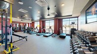 Located at 120 Matheson Ave in Charlotte, NC, this fitness center offers a spacious, single-floor exercise room designed to accommodate a comprehensive range of workout routines. The interior is defined by bold deep-red accent walls contrasted against a dark, textured ceiling with recessed and pendant lighting, creating an energizing yet focused atmosphere.

Large floor-to-ceiling windows line one side of the room, flooding the space with natural light and providing open views of the surrounding area. The flooring consists of durable, neutral-toned carpet suited for high-traffic athletic use.

The cardio zone features a row of TRUE-brand treadmills and elliptical machines, all equipped with individual digital displays. Strength training is well-represented with a full dumbbell rack spanning a range of weights, displayed beneath a wall-mounted flat-screen television. A multi-functional power rack supports TRX suspension systems, medicine balls, and battle ropes, offering versatile functional training options. Additional cable and weight machines round out the strength equipment selection.

Full-length mirrors are strategically placed to enhance the sense of space and support proper form during workouts. The layout is open and thoughtfully organized, ensuring ample room to move between stations. Overall, this fitness center is a fully equipped, modern facility built to support diverse training needs.
