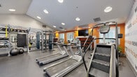 Located at 24080 Amador St in Hayward, California, this fitness center is a thoughtfully designed, single-floor facility that offers a comprehensive workout environment. The space is organized to accommodate a wide range of exercise needs, with distinct zones for cardio and strength training seamlessly integrated into an open floor plan.

The cardio area is outfitted with multiple Matrix-brand treadmills, elliptical trainers, a stair climber, and a recumbent bike, all arranged to maximize usability. A wall-mounted flat-screen TV adds convenience for those looking to stay entertained during workouts. The strength training section features a multi-functional cable machine, TRX suspension system, a full dumbbell rack, and a dedicated free weights area lined with floor-to-ceiling mirrors to support proper form.

Vibrant orange accent walls and an expansive motivational typography mural — displaying fitness-related words and phrases in bold orange and gray text — energize the atmosphere and reinforce a wellness-focused environment. The durable rubber flooring throughout provides both safety and comfort. Recessed ceiling lighting ensures the space is well-lit, while windows and glass double doors open directly to an outdoor courtyard, allowing natural light to filter in. The fitness center combines functionality with visual energy, making it an inviting space for daily exercise.
