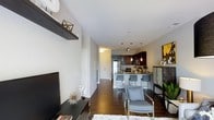 This well-appointed single-floor residence showcases a thoughtfully designed open-concept layout that seamlessly connects the living and kitchen areas. Rich dark hardwood flooring runs throughout, anchoring the space with warmth and elegance. The living area features a sleek floating wall shelf, a wall-mounted flat-screen TV, and a plush area rug, creating an inviting yet refined atmosphere. A modern accent chair and carefully curated décor add personality and style to the space. The kitchen is a standout, boasting rich dark cabinetry, pendant lighting that casts a warm glow, stainless steel appliances, and a granite-topped breakfast bar with contemporary bar seating — ideal for casual dining or entertaining. Recessed lighting throughout ensures the space is well-lit, complementing the neutral, light gray wall tones that keep the interior feeling bright and open. The open corridor leading to the entryway provides a clear sense of the unit's generous depth, while the overall design balances modern aesthetics with functional living. Every detail, from the hardware to the fixtures, reflects a commitment to contemporary style and quality craftsmanship.