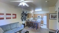 This single-floor space offers an open-concept layout that integrates the living area and kitchen into one cohesive, functional environment. The living room is a comfortable and inviting space, featuring a ceiling fan with light fixture that provides both airflow and ambient lighting. Neutral-toned walls and carpeted flooring create a warm, welcoming atmosphere throughout.

The kitchen is well-appointed with light wood cabinetry that extends to the upper walls, providing ample storage. A full suite of white appliances — including a refrigerator, electric range, and over-the-range microwave — lines the back wall. The kitchen flows naturally into the living space by way of a peninsula-style breakfast bar with a countertop overhang, accommodating multiple bar stools and serving as an ideal spot for casual dining or entertaining.

Tile flooring in the kitchen offers a practical contrast to the carpeted living area, clearly defining each zone while maintaining an open feel. The overall design emphasizes functionality and ease of living, with a layout that encourages social interaction between the kitchen and living spaces. Natural decorative elements, including a large indoor plant, add a touch of warmth and life to the interior.