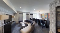 This thoughtfully designed media room offers a multifunctional entertainment experience across a single, cohesive floor. The space is anchored by a large flat-screen television mounted against a striking textured wallcovering that extends throughout the room, creating a unified and sophisticated visual backdrop. In front of the screen, a generously proportioned curved sectional sofa in warm neutral tones invites relaxed viewing, complemented by unique metallic tree-stump side tables that add an organic, artistic contrast to the contemporary interior.

Recessed lighting throughout the ceiling provides consistent illumination, while floor lamps flanking the TV wall contribute warmth to the media viewing area. A dedicated game zone features a shuffleboard table alongside additional seating, offering an interactive leisure option. Adjacent to this, a long communal dining or gathering table is surrounded by upholstered chairs beneath an industrial-style globe chandelier, making it ideal for group socializing.

Large windows dressed with two-tone curtains allow natural light to filter into the space, enhancing its open and inviting ambiance. Vibrant, colorful artwork depicting bottles is displayed prominently on one wall, providing a bold focal point that enlivens the otherwise neutral palette. Dark hardwood flooring in the gaming and dining zones transitions to plush carpeting in the lounge area, further delineating the room's distinct functional areas.