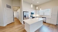 This single-story residence spans 1,190 square feet and offers 5 bedrooms, 2 bathrooms, and an open-concept kitchen and living area. Light hardwood flooring runs consistently throughout, creating a clean, cohesive aesthetic across all rooms. The kitchen is a standout feature, boasting tall white shaker-style cabinetry extending to the ceiling, a central island with white quartz countertops, stainless steel appliances including a refrigerator, range, microwave, and dishwasher, and a undermount sink beneath a window. Pendant lighting and recessed ceiling lights illuminate the space, which benefits from soaring ceilings that enhance its open feel. The adjoining living area features multiple large windows with white blinds, flooding the room with abundant natural light. The five bedrooms are finished with neutral-toned walls, clean white panel doors with modern hardware, and ceiling fans. The primary bedroom measures 140 square feet. The two bathrooms are finished in a contemporary style: one features a walk-in glass-enclosed shower with large-format grey tile, and the other includes a soaking tub with a tiled surround. Both bathrooms include white vanities and wood-look flooring, maintaining the home's consistent, modern design language.