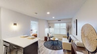 This single-floor residence offers a thoughtfully designed open-concept layout that seamlessly connects the kitchen, dining, and living areas. The kitchen is outfitted with rich dark-wood cabinetry, stainless steel appliances including a refrigerator and dishwasher, a undermount sink, and a central island with a white quartz countertop that doubles as a breakfast bar with barstool seating. Light wood-style flooring runs throughout the main living spaces, complementing the neutral color palette and adding warmth to the interior. The living area features a large window dressed with horizontal blinds, allowing ample natural light to fill the room, and offers comfortable space for seating and relaxation. The bedroom is a serene retreat with wide windows providing natural light, a closet, and light wood flooring consistent with the rest of the unit. The full bathroom includes a tiled tub-shower combination with floor-to-ceiling tile surround and clean white fixtures. A particularly functional feature is the in-unit laundry area, which includes a front-loading washer, a stone countertop vanity with an undermount sink, and glass wall-mounted storage shelves. Recessed lighting and track lighting are found throughout, enhancing the bright and modern ambiance of the space.