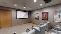 This dedicated theater room delivers an immersive cinematic experience within a thoughtfully designed, self-contained space. The room centers on a large wall-mounted projection screen paired with a ceiling-mounted projector, providing a sharp, theater-quality viewing experience. Recessed lighting throughout the ceiling offers adjustable ambiance, while flush-mounted in-ceiling speakers ensure comprehensive surround sound coverage for an authentic audio experience.

Seating consists of multiple rows of plush, upholstered armchairs finished in a soft patterned fabric, each equipped with built-in cup holders and foldable ottomans for maximum comfort during extended viewing sessions. The layout accommodates a generous number of guests, making it well suited for group screenings. Neutral taupe walls and warm wood-toned baseboards lend a polished, finished aesthetic to the room, while textured carpet flooring adds warmth and acoustic absorption.

Adding to the cinematic atmosphere, framed classic movie posters — including Titanic, Pirates of the Caribbean, and The Karate Kid — adorn the walls, reinforcing the theater's character and charm. A solid wood entry door provides privacy and sound separation from adjacent spaces. Discreet control panels near the entry allow for convenient management of the room's audiovisual and environmental systems, completing this well-appointed, fully functional private theater.