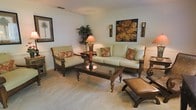 This single-story space showcases a thoughtfully designed living area that blends comfort with tropical-inspired elegance. The living room is furnished with a plush, light sage-green sofa and a matching armchair, both crafted from dark wood frames and accented with rust and floral patterned throw pillows that add a warm, inviting contrast. A substantial dark wood coffee table with a glass top serves as the room's centerpiece, complemented by coordinating side tables and decorative accent pieces, including a carved wooden ottoman. The neutral, light-toned carpet throughout the living space adds softness underfoot and enhances the room's airy feel. Walls are painted in a clean, soft white and feature subtle wainscoting details, providing a refined architectural element. Décor includes a bold botanical canvas print, a framed palm tree artwork, and paired wall sconces that contribute to the tropical ambiance. A tall decorative plant adds a natural, lush element to the corner of the room. The overall atmosphere is warm, cohesive, and relaxed, reflecting a casual yet polished aesthetic well-suited for comfortable everyday living.