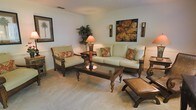 This single-story space showcases a thoughtfully designed living area that blends comfort with tropical-inspired elegance. The living room is furnished with a plush, light sage-green sofa and a matching armchair, both crafted from dark wood frames and accented with rust and floral patterned throw pillows that add a warm, inviting contrast. A substantial dark wood coffee table with a glass top serves as the room's centerpiece, complemented by coordinating side tables and decorative accent pieces, including a carved wooden ottoman. The neutral, light-toned carpet throughout the living space adds softness underfoot and enhances the room's airy feel. Walls are painted in a clean, soft white and feature subtle wainscoting details, providing a refined architectural element. Décor includes a bold botanical canvas print, a framed palm tree artwork, and paired wall sconces that contribute to the tropical ambiance. A tall decorative plant adds a natural, lush element to the corner of the room. The overall atmosphere is warm, cohesive, and relaxed, reflecting a casual yet polished aesthetic well-suited for comfortable everyday living.