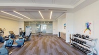 This fitness center presents a thoughtfully designed, single-level space that combines contemporary architecture with a comprehensive range of exercise equipment. The layout is open and spacious, with patterned carpet tiles in muted earth tones providing a practical yet polished foundation throughout. Recessed linear LED lighting runs across the ceiling, delivering bright, even illumination that complements the abundant natural light streaming in through floor-to-ceiling glass walls and large windows overlooking outdoor greenery.

The cardio zone features a well-curated selection of upright bikes, recumbent bikes, ellipticals, and treadmills arranged to maximize space and usability. Adjacent to this area, a dedicated strength training section houses a variety of cable and selectorized machines suited for full-body workouts. A clearly defined free weights area includes a multi-tiered dumbbell rack with a full range of weights, accompanied by a flat bench for targeted exercises.

A functional training corner provides stability balls, medicine balls on a vertical rack, and weight plates, supporting dynamic and core-focused workouts. Built-in cabinetry with light wood veneer along one wall offers convenient storage. Colorful sports-themed artwork decorates the white walls, adding an energetic character. Glass partition walls define a secondary exercise area, maintaining visual openness while creating distinct workout zones.