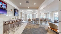 Located at 10051 Perimeter Pkwy in Charlotte, NC, this single-floor communal space functions as a stylish clubhouse or leasing lounge designed to accommodate both social gatherings and relaxed individual use. The open floor plan is thoughtfully organized into distinct seating zones, each with its own character while maintaining a cohesive contemporary aesthetic throughout.

Floor-to-ceiling windows along multiple walls flood the interior with natural light and offer outward views of the surrounding landscape. Sheer white drapery frames these expansive window walls, softening the light and adding an elegant touch to the neutral color palette of warm beiges, grays, and whites.

A bar counter area with stylish stools and a wall-mounted television anchors one side of the space, providing a social hub. Adjacent to it, high-top communal tables paired with patterned upholstered chairs create a collaborative dining or meeting area. Overhead, geometric linear pendant fixtures provide focused task lighting, complementing the recessed ceiling lights throughout.

Transitioning toward the lounge zones, curved upholstered chairs, rounded ottomans, and low-profile coffee tables are arranged atop layered area rugs, encouraging conversation and relaxation. Decorative wall art and accent lighting complete the refined interior, while the articulated tray ceilings with contrasting tones add architectural depth to the overall design.