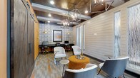 Located at 45 Lullwater Dr in Richmond Hill, Georgia, this single-floor community space blends contemporary design with warm, inviting character. The interior showcases a thoughtfully curated aesthetic, highlighted by a coffered ceiling with rich wood-toned paneling and recessed lighting that creates a polished, layered atmosphere. Statement geometric pendant chandeliers serve as focal points, casting warm light throughout the room and adding an industrial-chic flair to the space.

The lounge area is furnished with modern accent chairs and an upholstered ottoman in a bold amber tone, paired with sleek side tables — ideal for casual gatherings or relaxed conversation. Shiplap wall paneling in soft white contrasts beautifully with a vibrant accent wall adorned in a bold, ornate patterned tile motif, adding visual depth and personality to the room.

A built-in business center lines one wall, featuring a long dark-toned desk with multiple workstations, gold pendant lighting overhead, and ample space for productivity. Large windows allow natural light to filter in, enhancing the open, airy feel of the space. The flooring features a multi-toned, patterned carpet with orange accents that ties the warm color palette together cohesively throughout the room.