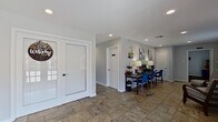 This single-story space is thoughtfully organized into several distinct functional zones, offering a well-rounded environment suited for both relaxation and productivity. The layout, as revealed by the floor plan, follows an L-shaped configuration that efficiently connects each area while maintaining a sense of separation between uses.

The entry area greets visitors through classic double French doors and leads into an open corridor finished with large-format stone-look tile flooring that carries throughout much of the space. Recessed lighting overhead keeps the atmosphere bright and clean against soft gray walls and crisp white trim.

A comfortable lounge area features large sliding glass doors that invite natural light and offer views of the outdoors. The seating arrangement, with upholstered wingback chairs and a central coffee table, creates a calm, welcoming retreat. An adjacent workspace includes a dark-finished desk and blue accent chairs beneath decorative wall art.

The kitchenette is well-appointed with dark cabinetry, stone-look countertops, a full-size refrigerator, microwave, and sink. A peninsula breakfast bar with rustic upholstered barstools provides casual seating and a social focal point.

A dedicated fitness area rounds out the space, equipped with various exercise machines, free weights, and treatment-style tables, making this space well-suited for health or wellness-oriented use.