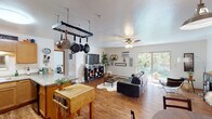 This single-floor residence offers a well-organized layout that balances open communal areas with private living spaces. The kitchen is equipped with warm honey-toned cabinetry, granite-look countertops, black appliances including an electric range and over-the-range microwave, and a double-basin sink. A pass-through window connects the kitchen to the adjacent living area, enhancing the open feel. A hanging pot rack above the kitchen island adds a functional, chef-inspired touch. The dining area sits just off the kitchen beneath a pendant light, featuring vaulted ceiling detail and wood-look flooring that extends throughout the main living spaces. The living room is spacious and bright, anchored by a ceiling fan and a wall-mounted air conditioning unit. Large sliding glass doors open directly to an outdoor patio and a lush, green yard surrounded by mature trees, flooding the room with natural light. The bedroom offers plush carpet, dual windows framing wooded views, and a ceiling fan. The bathroom includes a wide vanity with a large mirror and ample counter space. Overall, the home exudes a warm, casual ambiance.