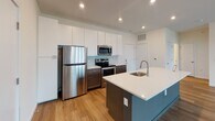 Located on West Broad Street in Richmond, Virginia, this single-floor unit offers a thoughtfully designed, open-concept layout with contemporary finishes throughout. Wood-look flooring flows consistently across the living areas and bedrooms, complementing the clean, neutral color palette that gives the space a bright and airy feel.

The kitchen serves as a central hub of the unit, featuring a teal-accented island with a white quartz countertop and undermount sink, tall white upper cabinetry, dark lower cabinets, and a herringbone-patterned tile backsplash. Stainless steel appliances, including a refrigerator, range, microwave, and dishwasher, complete the well-equipped space. Recessed lighting adds warmth overhead.

The open living area adjoins the kitchen seamlessly, with large windows fitted with white horizontal blinds that invite natural light while offering neighborhood views. The bedrooms continue the hardwood-style flooring and feature generous window banks. One bedroom includes a sliding barn door that opens to a walk-in closet with built-in shelving.

The bathrooms are finished with light tile flooring, dark wood vanities, white quartz countertops, and a tub-shower combination. Brushed nickel fixtures and grab bars add functional detail throughout.