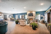 This single-floor community space is thoughtfully designed to offer a welcoming and functional environment across several distinct areas. The lounge area serves as a central gathering point, furnished with plush teal accent chairs arranged around a media console with a wall-mounted television, flanked by decorative planters and framed mirrors that add depth to the neutral-toned walls.

The kitchen is a standout feature, boasting rich dark wood cabinetry, a classic subway tile backsplash, and a full suite of stainless steel appliances including a side-by-side refrigerator, range, and microwave. A curved granite-topped island with pendant lighting and teal upholstered bar stools serves as both a workspace and casual gathering spot, with wood-look flooring grounding the space.

The dining area offers ample seating around a glass-topped table, set against a bold deep navy accent wall. French doors open to an outdoor view, allowing natural light to fill the room and creating a bright, inviting atmosphere.

The living area is anchored by a dramatic floor-to-ceiling stacked stone fireplace with a mounted television above, complemented by generous seating, a matching navy accent wall, and warm recessed lighting throughout. The combination of textures, colors, and thoughtful furnishings gives this space a comfortable yet refined character.