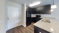 This single-floor residential unit offers a thoughtfully designed layout with a cohesive, modern aesthetic throughout. The kitchen serves as a functional centerpiece, featuring dark espresso cabinetry, granite-style countertops, and a full suite of black appliances including a refrigerator, range, and built-in microwave. Wood-look flooring runs through the kitchen, adding warmth and continuity to the space. The bathroom is well-equipped with a dark vanity cabinet topped with granite-style counters, an undermount sink, a large mirror with overhead lighting, wood-look flooring, and a shower-tub combination accented by a bold geometric curtain. The bedroom is carpeted for comfort and includes a ceiling fan with a light fixture, a window with blinds that allows natural light to filter in, and sufficient space for a full bedroom set and a study desk. The living area provides a relaxed setting with neutral walls and access to natural light through a sliding door or large window. Completing the unit is a dedicated laundry area housing a stacked Kenmore washer and dryer unit alongside a water heater, maximizing the use of the available utility space. Overall, the unit presents a practical and modern living environment with consistent finishes throughout.