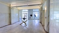 This elegant single-floor lobby showcases a refined blend of classical and contemporary design. The space is defined by its ornate crown molding and decorative plasterwork cornice that runs along the ceiling perimeter, lending a sense of historical grandeur. Large, floor-to-ceiling windows and glass-paneled entrance doors at the far end flood the interior with natural light, creating a bright and welcoming atmosphere.

The flooring is composed of light-toned marble or stone tiles, lending a polished, upscale character to the space. Marble wall panels frame the room on either side, reinforcing the classical aesthetic and adding visual depth. A sleek, modern glass-top table with a sculptural geometric base sits at the center of the lobby, providing a striking contemporary contrast to the traditional architectural surroundings. A floral arrangement atop the table adds a subtle organic touch.

The ceiling features exposed industrial-style conduit alongside the ornate molding, hinting at a thoughtfully preserved historic building adapted for modern use. Overall, the lobby exudes a sophisticated ambiance, balancing heritage architectural elements with clean, modern furnishings.