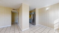 This single-floor unit offers a straightforward and functional layout with a consistent design language carried throughout. Light-colored ceramic tile flooring spans the entirety of the space, providing a cohesive and easy-to-maintain foundation in every room.

The living area features distinctive large-format tile accent walls in a warm cream tone, adding texture and a retro character to the space. A ceiling fan with a built-in light fixture provides both comfort and illumination, while a large window with blinds allows natural light to fill the room and offers views of the surrounding exterior.

The kitchen is efficiently laid out and equipped with stainless steel appliances, including a gas range and a full-size refrigerator. Gray cabinetry lines the upper walls, providing ample storage, and the tiled walls continue here, maintaining a uniform aesthetic. A central structural column partially separates the kitchen from the adjoining living and dining areas, creating a sense of defined zones without fully closing off the space.

The bathroom is finished with floor-to-ceiling tile work featuring a decorative mosaic border accent above the tub surround. A full-size soaking tub with an overhead shower, along with a standard toilet, complete the bathroom.

The entry area opens directly into the main living space, anchored by a solid white front door flanked by a wide window, welcoming natural light from the moment you step inside.