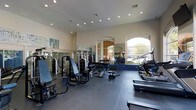This single-level fitness center offers a comprehensive workout environment designed to accommodate a wide range of exercise needs. The space is generously sized, with elevated ceilings and recessed lighting that create a bright, open atmosphere. Large arched windows along one wall allow abundant natural light to flood the interior, offering views of the surrounding outdoor area and enhancing the overall sense of openness.

The flooring features a dark rubberized surface in the main workout zone, providing cushioning and traction for both strength and cardio activities. Floor-to-ceiling mirrors line the walls, visually expanding the space while allowing users to monitor their form during exercise.

The equipment layout is thoughtfully organized, with a dedicated cardio section along one side housing treadmills and elliptical machines. The strength training area features a variety of selectorized machines, benches, and a dumbbell rack with a full weight selection. Motivational fitness-themed artwork adorns the walls, contributing to an energetic and encouraging atmosphere. The overall design balances functionality with a clean, modern aesthetic suited for daily athletic use.