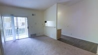 This single-floor space offers a straightforward, functional layout centered around an open living area finished with neutral wall-to-wall carpeting. The room is defined by clean, cream-colored walls and a textured ceiling, giving the space a simple and understated character. A notable architectural feature is the pass-through window cut into a partial dividing wall, which connects the living area to an adjacent room—likely a kitchen—while maintaining a sense of openness. A small wooden cabinet base anchors the base of this dividing wall, adding a touch of warmth to the otherwise neutral palette. Natural light enters through a glass-paneled door on the far wall, which opens to what appears to be a balcony or patio with a railing visible beyond. A floor-level heating vent is positioned near this door, ensuring comfort throughout the space. The layout allows for flexible furniture arrangement and provides a clear sense of flow between areas. Overhead HVAC vents are integrated into the ceiling, supporting year-round climate control. Overall, the space presents a clean, move-in-ready environment with practical features and a calm, neutral aesthetic.