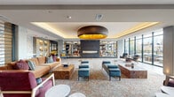 This generously scaled amenity lounge serves as a striking communal centerpiece, thoughtfully designed to balance elegance with comfort. The main interior space is anchored by a dramatic circular pendant light with a warm gold interior, suspended above a coffered ceiling with soft cove lighting that lends a refined, hotel-like ambiance throughout. A sleek, floor-to-ceiling linear fireplace serves as a focal point, flanked by expansive built-in shelving units displaying curated décor and books, adding both visual depth and warmth to the room.

Multiple distinct seating areas are arranged across the open floor plan, furnished with a carefully curated mix of mid-century modern and contemporary pieces — from tufted ottomans and burl wood coffee tables to leather accent chairs and plush sectional sofas. Patterned area rugs define each zone, while large floor-to-ceiling windows flood the interior with natural light and frame views of the outdoor patio beyond.

The patio is a standout feature, offering a spacious terrace with dining and lounge seating, framed by a railing that opens to panoramic views of the surrounding urban landscape and treetops. The entry corridor features polished tile flooring, curated greenery, and clean architectural lines, establishing an immediate sense of quality from the moment of arrival. Gold-accented elevator doors complete the upscale aesthetic.
