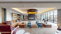 This generously scaled amenity lounge serves as a striking communal centerpiece, thoughtfully designed to balance elegance with comfort. The main interior space is anchored by a dramatic circular pendant light with a warm gold interior, suspended above a coffered ceiling with soft cove lighting that lends a refined, hotel-like ambiance throughout. A sleek, floor-to-ceiling linear fireplace serves as a focal point, flanked by expansive built-in shelving units displaying curated décor and books, adding both visual depth and warmth to the room.

Multiple distinct seating areas are arranged across the open floor plan, furnished with a carefully curated mix of mid-century modern and contemporary pieces — from tufted ottomans and burl wood coffee tables to leather accent chairs and plush sectional sofas. Patterned area rugs define each zone, while large floor-to-ceiling windows flood the interior with natural light and frame views of the outdoor patio beyond.

The patio is a standout feature, offering a spacious terrace with dining and lounge seating, framed by a railing that opens to panoramic views of the surrounding urban landscape and treetops. The entry corridor features polished tile flooring, curated greenery, and clean architectural lines, establishing an immediate sense of quality from the moment of arrival. Gold-accented elevator doors complete the upscale aesthetic.
