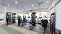 This fitness center is designed to support a full range of workout routines in a clean, well-organized environment. The space is outfitted with multiple treadmills equipped with individual digital displays, elliptical trainers, a stationary recumbent bike, and a multi-station cable and weight machine, providing users with both cardiovascular and strength-training options. A dedicated free-weight and resistance machine area rounds out the equipment selection.

The interior features a light-colored striped floor with alternating neutral tones, contributing to a clean and structured aesthetic. Ceiling fans are suspended from above, complementing the recessed lighting to maintain comfortable airflow and consistent illumination throughout the space. Large windows line the walls, allowing natural light to fill the room and offering an open, airy atmosphere.

A mirrored wall on one side visually expands the space while allowing users to monitor their form during workouts. The white walls and bright overhead lighting create a fresh, energizing environment well-suited for regular fitness use. The layout is thoughtfully arranged to allow clear circulation between equipment stations.