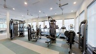 This fitness center is designed to support a full range of workout routines in a clean, well-organized environment. The space is outfitted with multiple treadmills equipped with individual digital displays, elliptical trainers, a stationary recumbent bike, and a multi-station cable and weight machine, providing users with both cardiovascular and strength-training options. A dedicated free-weight and resistance machine area rounds out the equipment selection.

The interior features a light-colored striped floor with alternating neutral tones, contributing to a clean and structured aesthetic. Ceiling fans are suspended from above, complementing the recessed lighting to maintain comfortable airflow and consistent illumination throughout the space. Large windows line the walls, allowing natural light to fill the room and offering an open, airy atmosphere.

A mirrored wall on one side visually expands the space while allowing users to monitor their form during workouts. The white walls and bright overhead lighting create a fresh, energizing environment well-suited for regular fitness use. The layout is thoughtfully arranged to allow clear circulation between equipment stations.