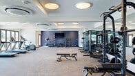 This fitness center offers a thoughtfully designed, single-floor layout that accommodates a comprehensive range of workout needs. The space is divided into distinct functional zones, each outfitted with professional-grade equipment. The cardio area lines a wall of large, floor-to-ceiling glass doors that flood the room with natural light and provide open views to the outdoors, creating an energizing environment for treadmills, ellipticals, stair climbers, and stationary bikes. The strength training zone features a robust selection of free weights, including tiered dumbbell racks, barbells, and kettlebells, alongside cable machines, a power rack with an adjustable bench, and multiple multi-functional cable stations. A wall of mirrors extends across this area, visually expanding the space and supporting proper form. The accent walls, finished in a deep navy blue, add a modern, motivating aesthetic throughout the gym. Ceiling-mounted circular light fixtures and a centrally mounted flat-screen television enhance the ambiance and functionality. A dedicated stretching and open-floor area provides ample room for dynamic movement and flexibility training. The carpeted flooring with rubberized sections in key workout areas completes this well-appointed fitness facility.
