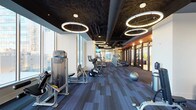 This contemporary fitness center offers a thoughtfully designed workout environment that balances functionality with sleek, modern aesthetics. The space is anchored by a dramatic open ceiling finished in dark tones, from which a series of large circular ring pendant lights hang at varying heights, creating a bold and energetic ambiance throughout the room. Floor-to-ceiling windows line one side of the space, flooding the interior with abundant natural light and providing open views of the surrounding area — an inspiring backdrop for any workout. The flooring features a durable, patterned carpet tile in cool gray tones, offering both comfort underfoot and a polished, professional look. The equipment layout is organized and spacious, with a dedicated cardio zone featuring treadmills, stationary bikes, and elliptical machines from leading brands such as Precor, all arranged to maximize flow and accessibility. A strength and resistance training area complements the cardio section, rounding out the facility's offerings. Toward the rear of the space, glass-paneled doors open to an adjacent area, extending the sense of depth and connectivity. Stability balls and additional accessories are thoughtfully placed throughout, making this fitness center a comprehensive and inviting space for a full range of exercise routines.