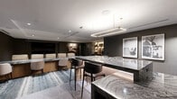 Located in the prestigious Buckhead district of Atlanta, this meticulously designed one-bedroom space delivers a compelling blend of luxury and functionality across a single thoughtfully planned floor. The living area commands immediate attention with its home theater setup, where a row of plush cream leather reclining seats faces a large-format screen set into a dramatic dark accent wall — an immersive cinematic experience within a residential setting. Adjacent to the theater zone, a stunning marble island anchored beneath a sleek rectangular pendant light serves as a central gathering point, its bold, sweeping veining lending an unmistakable artistic quality to the space. Large-format tile flooring transitions seamlessly into a textured blue-grey area rug, defining the lounge and dining zones. A sculptural marble dining table with curved upholstered chairs echoes the island's material palette, reinforcing a cohesive design language. Along one wall, backlit built-in shelving showcases curated decorative objects, adding warmth and dimension to the otherwise moody, dark-toned interior. The deep charcoal walls and recessed lighting throughout create an intimate, refined ambiance. The generously proportioned primary bedroom rounds out this elegantly appointed space, completing a residence of exceptional style and character.