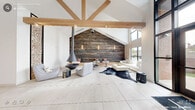 This expansive, single-floor space is a masterclass in contemporary design, blending rustic and modern elements to create a welcoming and visually striking environment. The soaring vaulted ceiling, framed by prominent exposed timber beams, immediately commands attention upon entry, lending the space a dramatic sense of height and openness. Large-format light-toned floor tiles stretch across the entire floor, enhancing the bright and airy atmosphere.

The layout is thoughtfully organized into distinct functional zones. A lounge area anchored by a sleek, suspended conical fireplace and a cowhide area rug provides an intimate gathering space, complemented by contemporary seating arrangements. A dramatic reclaimed wood accent wall, illuminated by vertical linear sconces, serves as a striking focal point, with built-in bench seating and small bistro tables nestled against it.

A dining and collaborative area features wooden tables and mixed seating, positioned near a decorative floor-to-ceiling stacked log display. Toward the entrance, a reception or leasing area is defined by dark-paneled walls and modern pendant lighting. Floor-to-ceiling windows and glass double doors at the entry flood the interior with natural light and offer views of the surrounding greenery, reinforcing the connection between the interior and the outdoors.
