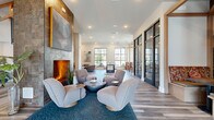 Located at 6026 Station Crossing Ave in Charlotte, NC, this thoughtfully designed community living space offers a generous single-floor layout that blends contemporary aesthetics with warm, inviting elements. The main living area serves as the heart of the space, anchored by a striking floor-to-ceiling tile fireplace wall adorned with large-scale artwork, surrounded by curved, modern lounge chairs atop a plush area rug. Wide-plank wood-look flooring flows throughout, unifying the various zones of the space. Natural light pours in through an abundance of black-framed windows and glass doors, creating a bright and open atmosphere with views to the outdoors. A welcoming entry foyer features a chandelier pendant light and additional lounge seating. Built-in wood-framed banquette nooks, accented by a dramatic mural of the Charlotte skyline, provide intimate gathering spots with pendant lighting overhead. Glass-partitioned rooms along the perimeter offer private or semi-private spaces for focused use. A dedicated refreshment bar showcases navy cabinetry, a white subway tile backsplash, and soaring ceilings with exposed wood beam accents and oversized pendant fixtures. The overall ambiance is modern, polished, and community-oriented — a sophisticated space designed for both socializing and productivity.