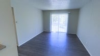 This single-floor space offers a clean, straightforward layout centered around a bright and open living area. The room is finished with dark hardwood-style flooring that contrasts neatly with the light gray, neutral-toned walls, creating a balanced and understated aesthetic throughout. A textured popcorn ceiling adds a classic touch overhead.

The most prominent feature of the living area is a large sliding glass door dressed with white vertical blinds, which serves as the primary source of natural light. The door allows an abundance of daylight to filter into the room, brightening the space and offering a view of the greenery outside. The sliding door also provides direct access to an outdoor area, enhancing the sense of openness and connection to the exterior environment.

White baseboards line the perimeter of the room, providing a clean border between the flooring and walls. The space is unfurnished, allowing for a clear view of its full dimensions and layout potential. Overall, the living area presents a neutral, move-in-ready environment with functional design elements and a light-filled atmosphere suited to a variety of interior arrangements.