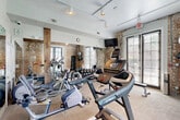 Located at 427 Josephine St in New Orleans' historic Garden District, this fitness center blends classic architectural character with modern workout amenities. The space is defined by striking exposed brick walls accented by rustic wooden beams, creating a warm, industrial aesthetic that reflects the building's heritage. Large windows fitted with blinds line the walls, flooding the room with natural light while offering views of the surrounding outdoor area, including a glimpse of a pool beyond the French doors. Track lighting mounted on the ceiling provides additional illumination throughout. The exercise room is well-equipped for a full workout experience, featuring treadmills, elliptical machines, a stationary bike, and a multi-function weight bench. A full dumbbell rack stocked with a wide range of weights caters to strength training needs. Wall-mounted accessories include a yoga mat, stability ball, and resistance tools for versatile training options. A large mirror spans one wall, enhancing the sense of space and allowing users to monitor their form. The light wood-toned flooring is durable and functional, suitable for varied workout activities. Despite its compact footprint, the thoughtful layout and abundant equipment make this fitness center a highly functional and visually appealing space.