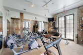Located at 427 Josephine St in New Orleans' historic Garden District, this fitness center blends classic architectural character with modern workout amenities. The space is defined by striking exposed brick walls accented by rustic wooden beams, creating a warm, industrial aesthetic that reflects the building's heritage. Large windows fitted with blinds line the walls, flooding the room with natural light while offering views of the surrounding outdoor area, including a glimpse of a pool beyond the French doors. Track lighting mounted on the ceiling provides additional illumination throughout. The exercise room is well-equipped for a full workout experience, featuring treadmills, elliptical machines, a stationary bike, and a multi-function weight bench. A full dumbbell rack stocked with a wide range of weights caters to strength training needs. Wall-mounted accessories include a yoga mat, stability ball, and resistance tools for versatile training options. A large mirror spans one wall, enhancing the sense of space and allowing users to monitor their form. The light wood-toned flooring is durable and functional, suitable for varied workout activities. Despite its compact footprint, the thoughtful layout and abundant equipment make this fitness center a highly functional and visually appealing space.