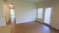 This single-story space in Denton offers a clean, functional layout with a bright and open living area. The main room is finished with warm-toned wood-style flooring that runs throughout, providing a cohesive and easy-to-maintain surface underfoot. Walls are painted in a soft, neutral cream tone that enhances the sense of openness and pairs well with the natural light that enters through multiple windows fitted with white horizontal blinds. The windows allow for adjustable light control while maintaining a tidy, uniform appearance. White baseboards frame the room, adding a crisp contrast to both the flooring and walls. A doorway visible from the main living area leads to an adjacent room, suggesting a connected and accessible floor plan. The overall ambiance is understated and versatile, providing a neutral canvas well-suited for a variety of furnishing styles. The space presents a straightforward, livable environment with practical finishes throughout.